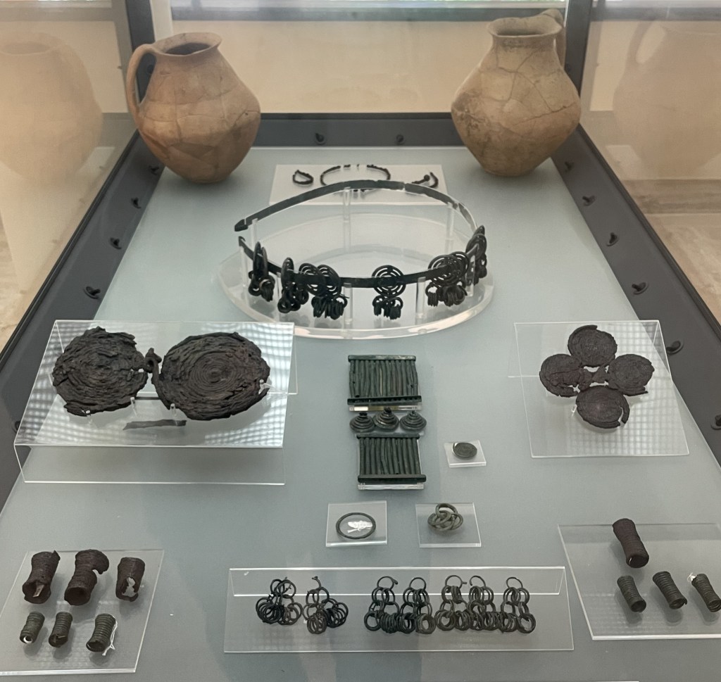 A museum display case filled with a number of metal items from a grave, including fibulae, two pots, rings, small cylindrical objects, and a headpiece.