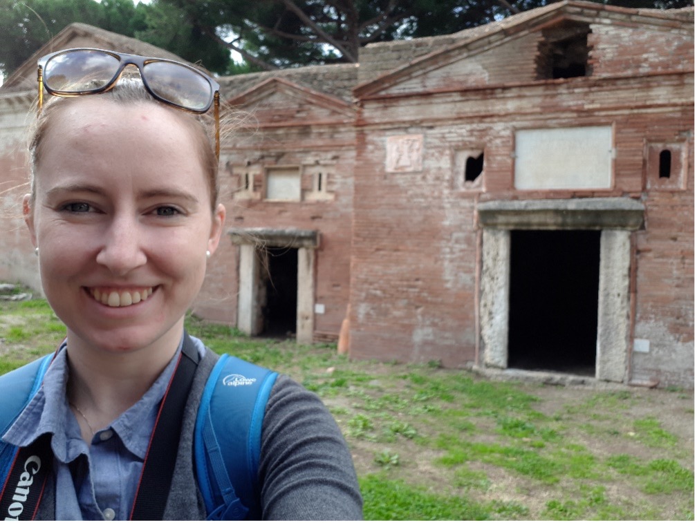 A white woman with blond hair. She has a pair of glasses on her head. She is standing in front of some house-like Roman tombs.