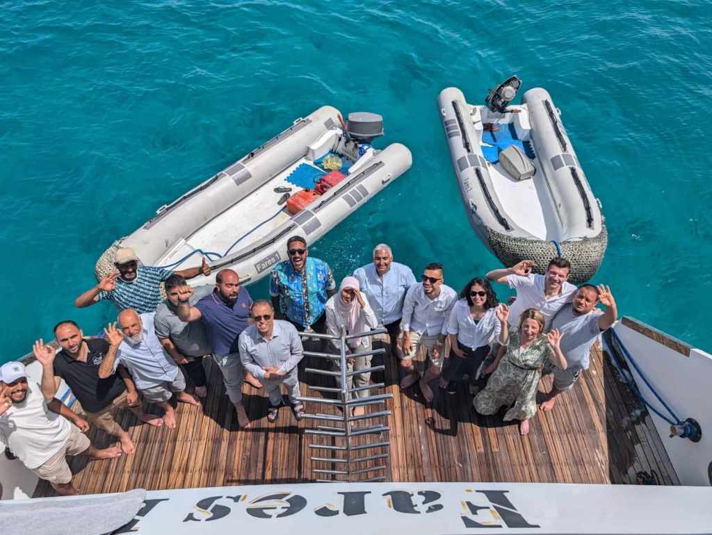 14 people stand at the stern of a pot looking up and waving to someone taking a photo from above. Two smaller motorized boats float in the water behind them.