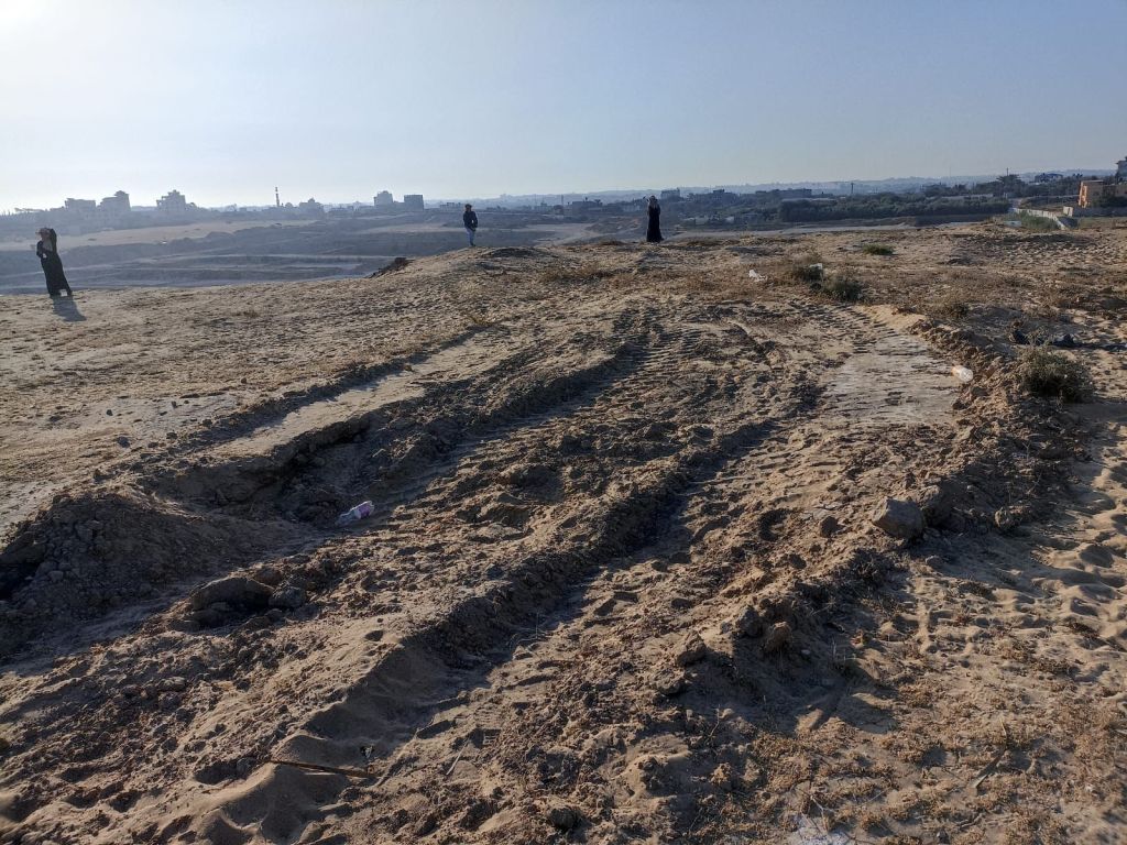 The image shows the site of Tell es Sakan; three people stand in the background, spaced out, engaged in survey work. Buildings can be seen in the further background.