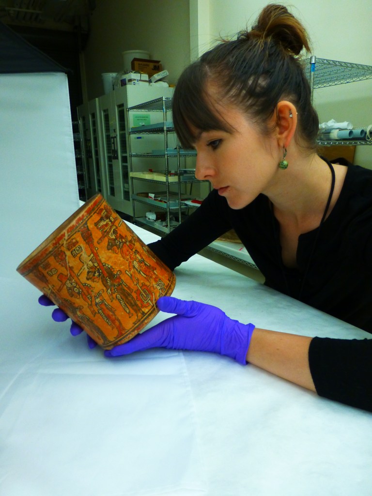 A white woman with brown hair holding a cylindrical ceramic vessel in her gloved hands.