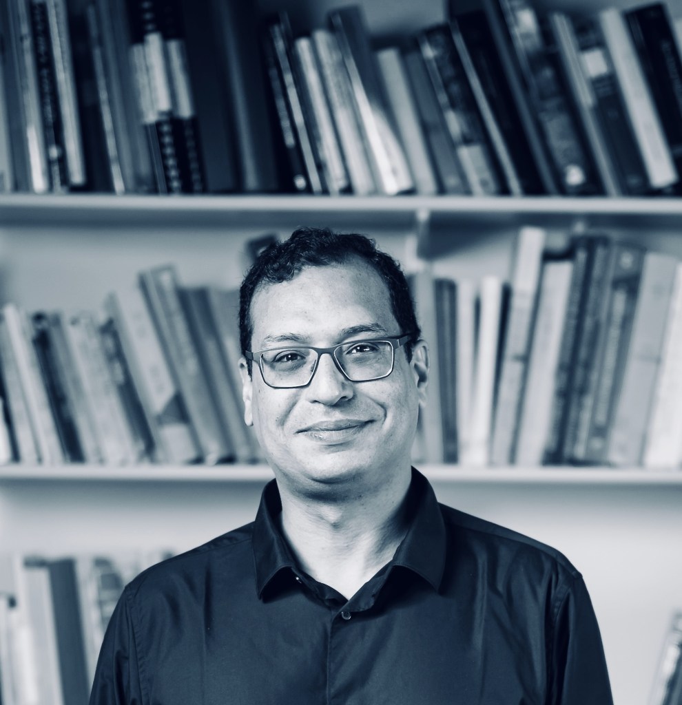 Black and white photo of a man with short dark hair and glasses, standing in front of a bookshelf.