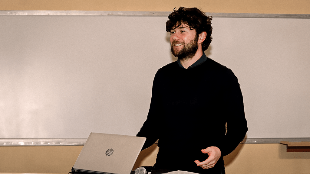 A white man with dark brown hair standing in front of a whiteboard and in front of a computer.