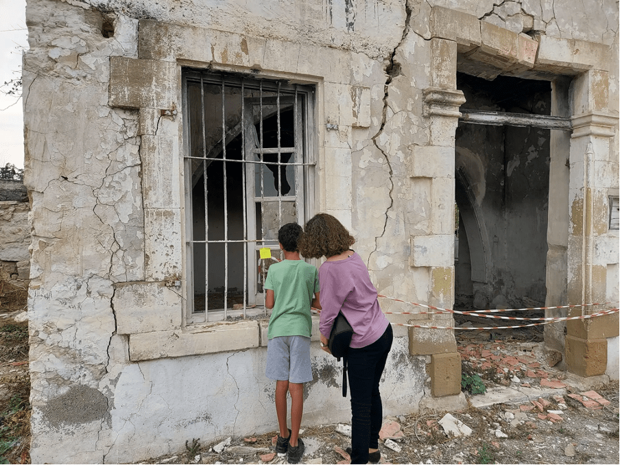 A boy wearing a green shirt and a woman wearing a mauve shirt standing in front of an abandoned building.