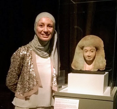 A woman wearing a grey headscarf, pink shirt, and a sparkly pink sweater, standing next to a bust of an Egyptian pharaoh in a museum case.