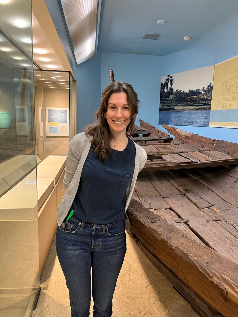 A white woman with shoulder-length brown hair standing in a museum between a display case and an ancient wooden boat. 