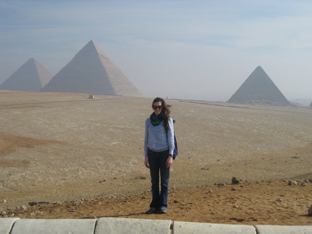 A white woman with shoulder-length brown hair standing in from of the pyramids at Giza.