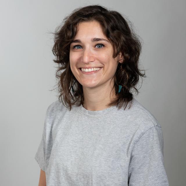 A white woman with shoulder length brown hair. She wears a grey t-shirt
