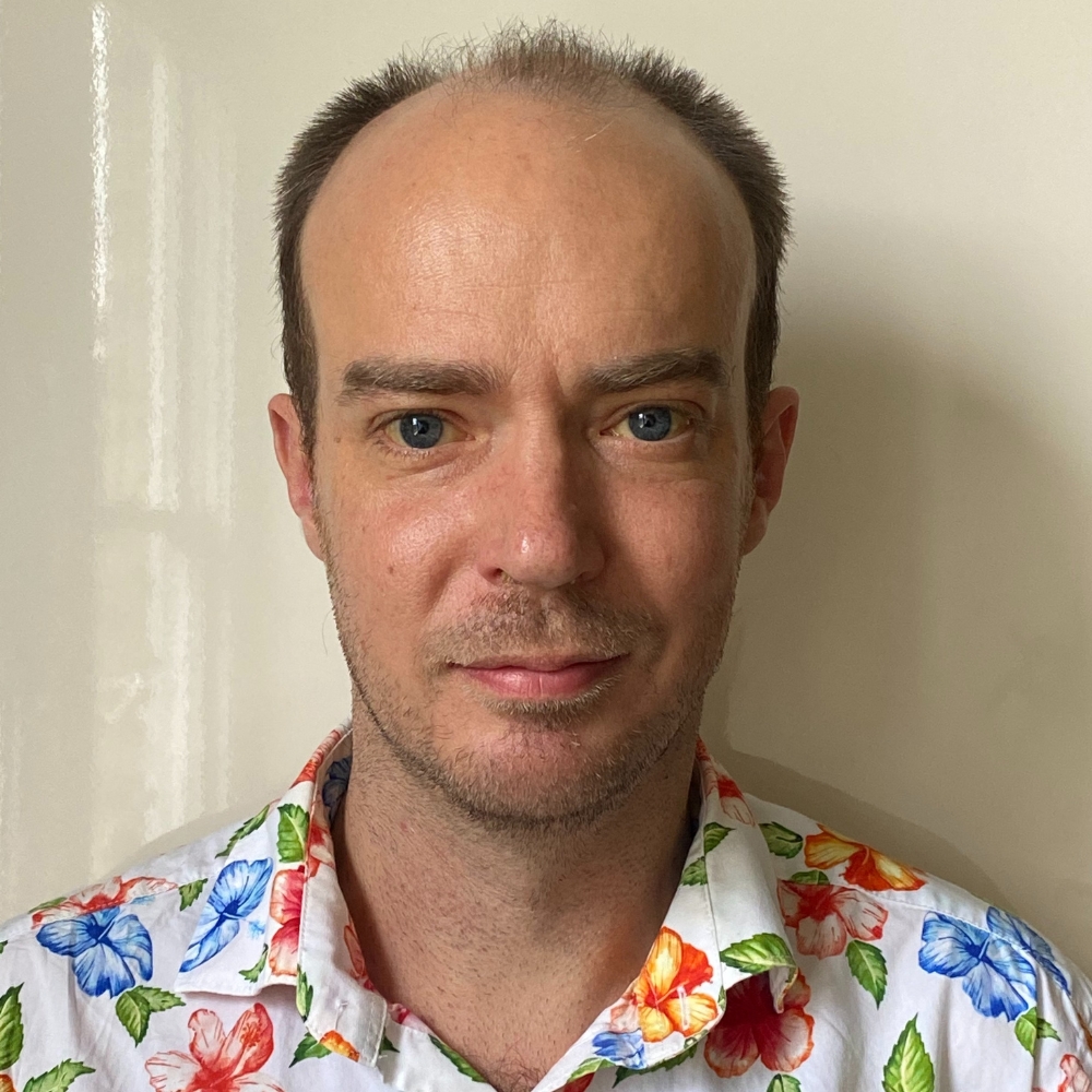 A white man with balding short brown hair. He wears a white shirt with flowers.