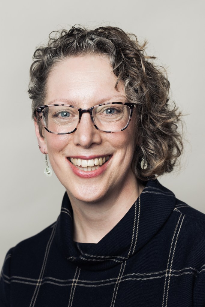 A white woman with short curly blond hair. She wears glasses and a black shirt. 