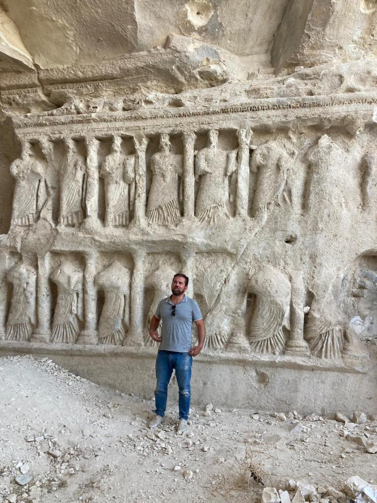 Man in a great shirt and jeans stands in front of a carved wall relief with two rows of nearly life-sized human figures.