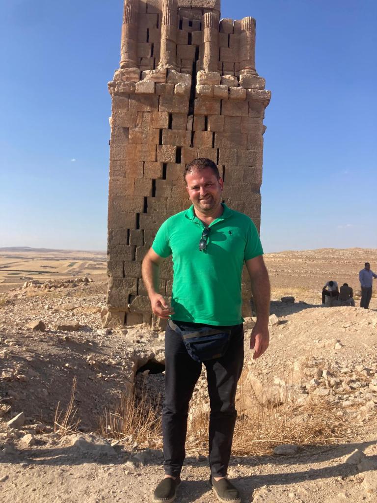 Man in a green shirt and jeans stands in front of a tower that shows a crack running down the centre.