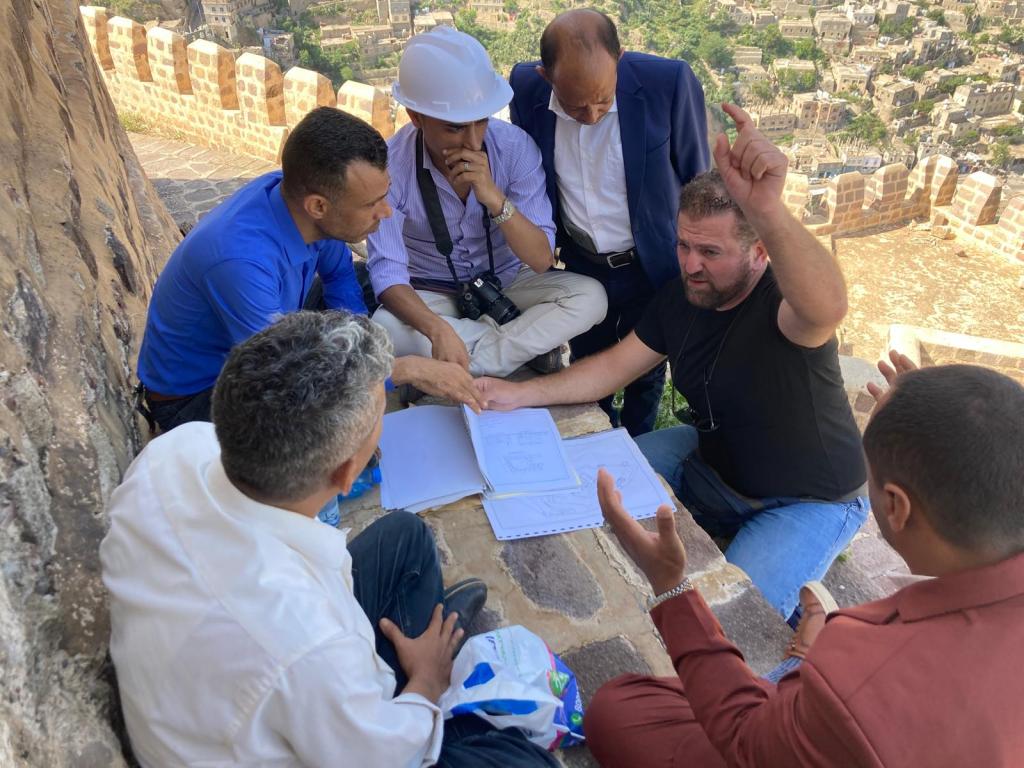 Group of people crouched down in conversation around a series of plans and documents. One man wears a construction helmet. The group is in the shade along the wall of a castle.
