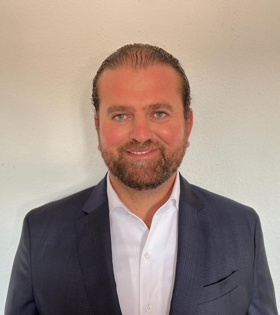 Headshot of a man with light hair and light eyes smiling at the camera. He wears a dark suit with a white shirt.
