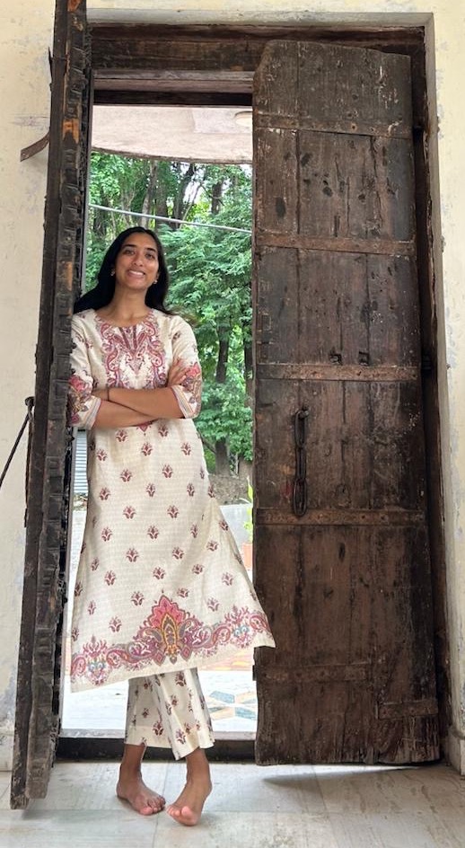 A young Indian woman with long black hair standing in front of a partially-opened wooden door. She wears a long white and pink dress over matching pants. 