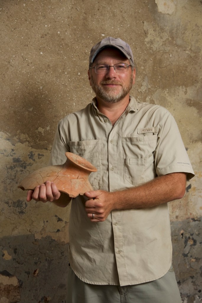 Photo of a white man with glasses, hold the upper part of a fragmented amphora.
