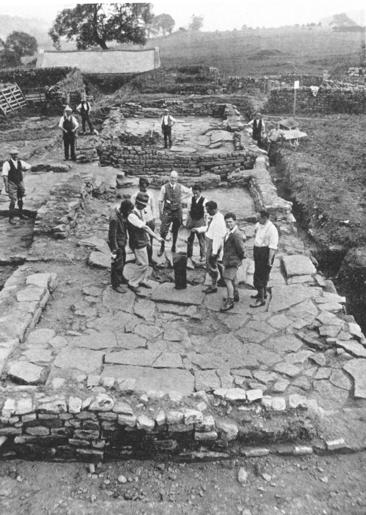 Black and white photo of several men standing around a feature in the middle of a stone paved room. Local workers stand in the background.