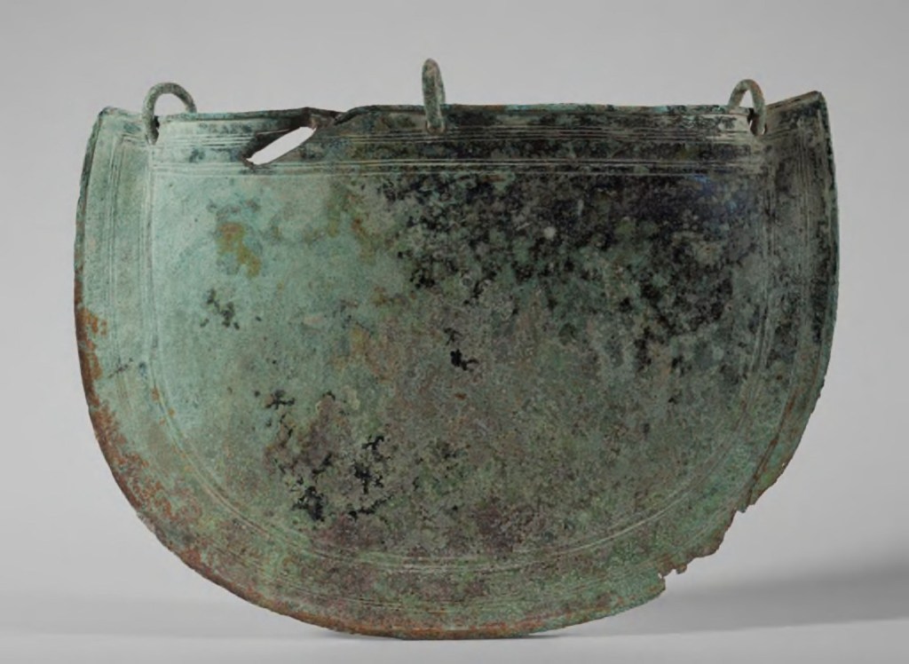 A semi-circular piece of bronze with three ring attachments and a hole (likely from a blade) near between the leftmost and centre bronze rings.