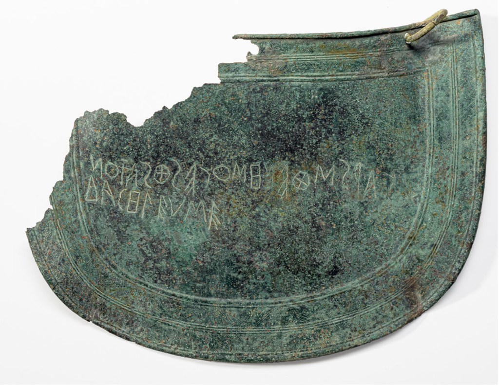 A broken piece of a semi-circular bronze groin guard, of which much of the upper left portion has disappeared. a singular bronze ring appears in the upper right of the guard. An Inscription is etched into the bronze.
