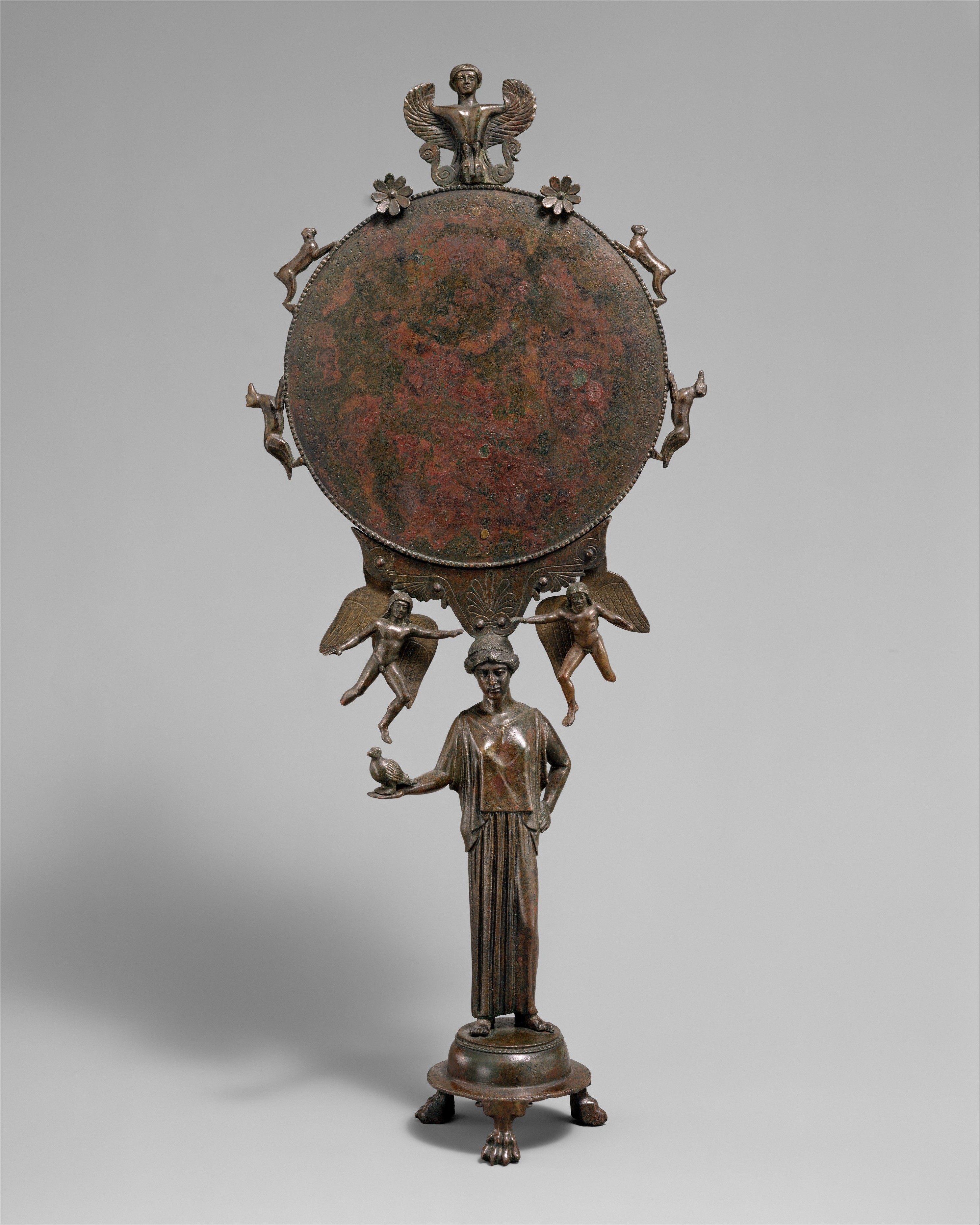 A bronze mirror in which the mirror is held up by a small female statuette. The circular mirror is surrounded by small winged figures, as well as two hounds and two hares. The top of the mirror has the bust of a siren.