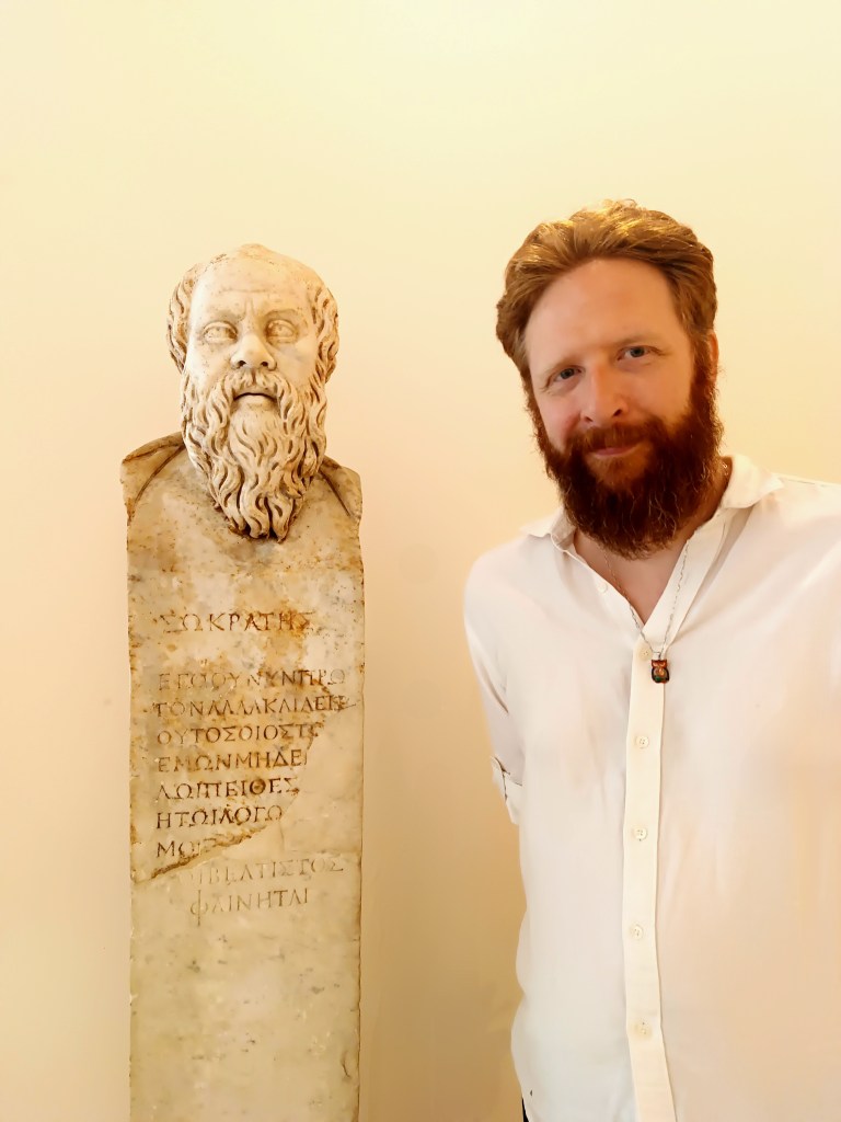 A white man with red hair and a red beard standing next to a white marble bust of Socrates