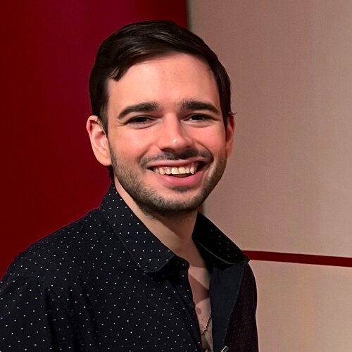 A headshot of a white male with short brown hair. He is wearing a black button up shirt with small white polka dots.