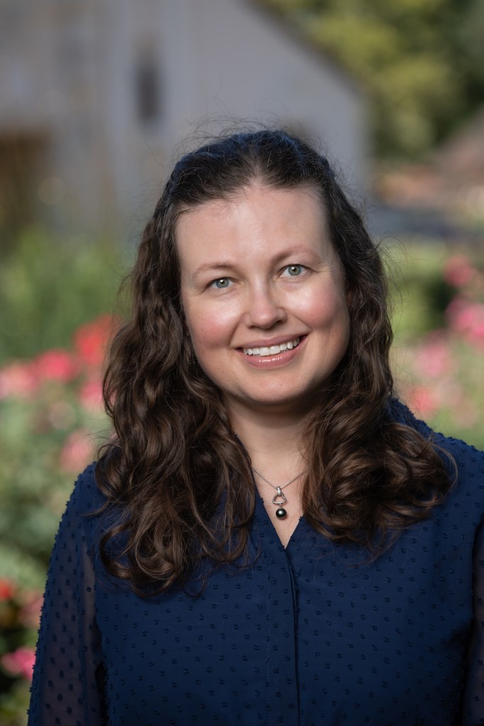 A white woman with shoulder-length brown hair. She wears a dark blue button-up shirt.