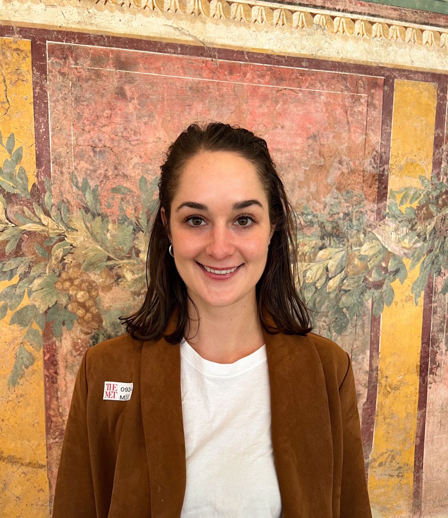 A Headshot of a white woman with dark brown hair. She wears a brown blazer with a white shirt underneath. She stands in front of a painted wall.