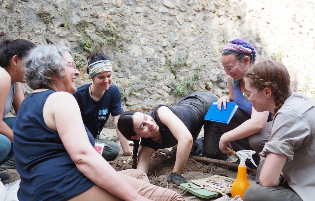 A group of four students sitting around a trench watching a white woman with dark brown hair excavation a small pit, as an older woman with grey hair smiles as she speaks to the students.