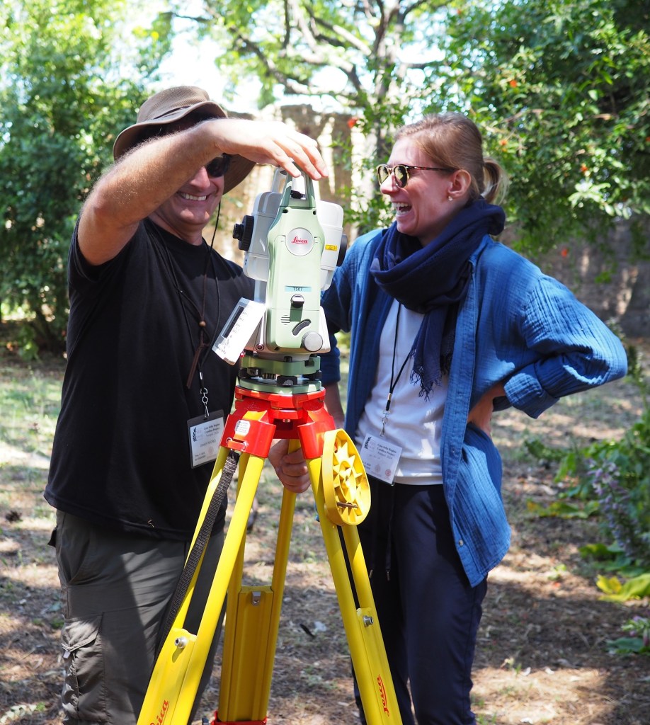 A male with a black shirt and wide-rimmed brown hat stands next to a woman with blond hair wearing a blue shirt and scarf. They both stand in front of a total station.