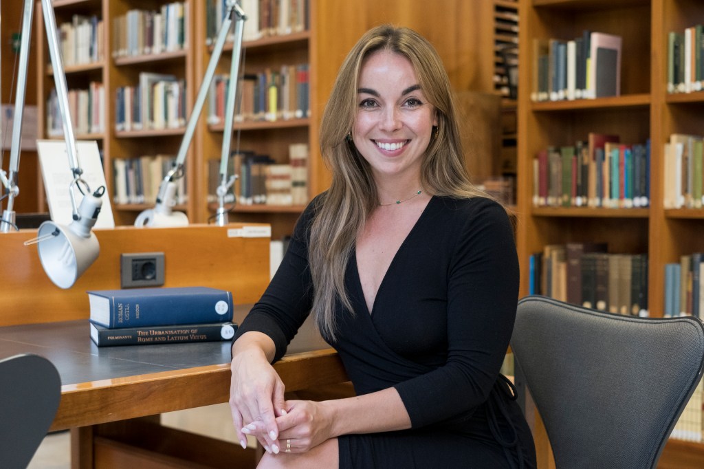 A blond woman wearing a black long-sleeved dress. She sits at a wooden table in a library. 