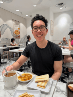 An Asian man with short dark hair and glasses, wearing a black t-shirt. He is sitting at a table in a restaurant with food in front of him.