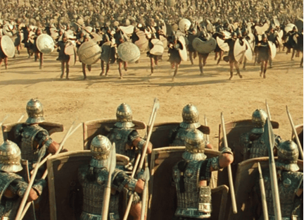A still image from the movie Troy, in which a group of soldiers runs towards a group of man standing in formation with their shields and spears up.