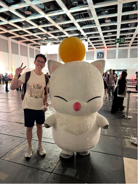 An Asian man holding up two fingers in a peace gesture, stands next to a large white character.