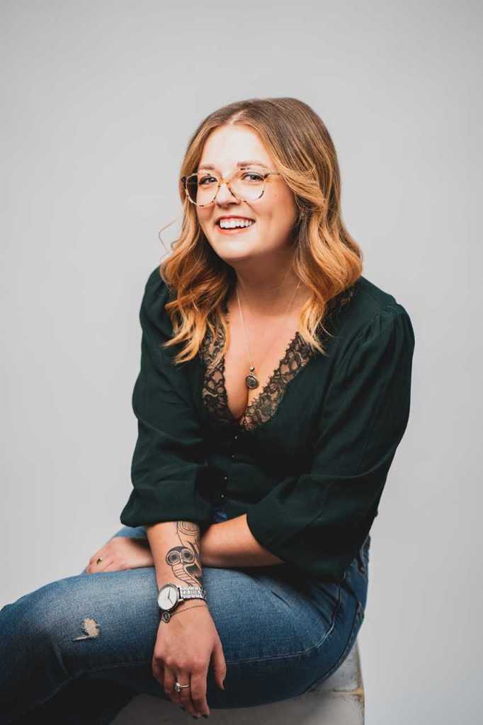 Image of Liv Albert, a white woman with blond hair, sitting on a stool. She wears a black shirt and jeans.