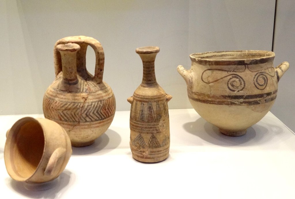 A museum display with a large drinking bowl with linear and spiral decoration to the right; next, to the left, is a thin jug iwht linear and geometric decoration; next is a spouted boar with linear and chevron decoration; to the left and slightly in front is a drinking cup tipped over