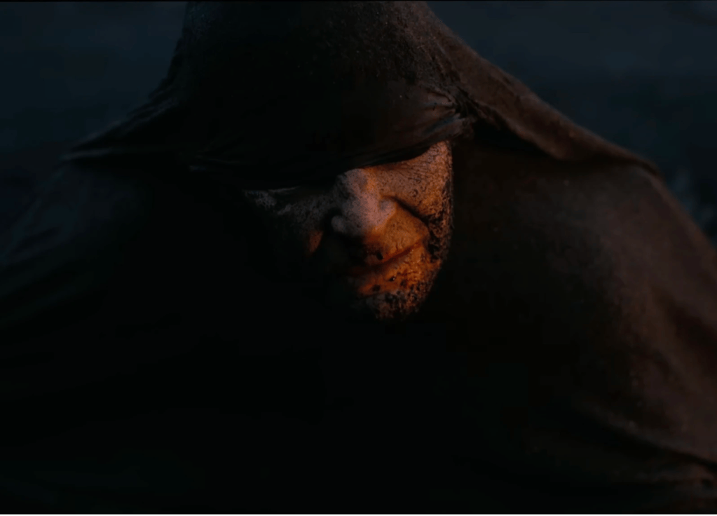 A dark image of a man with sand on his face, wearing a large black cloak that covers his eyes. 
