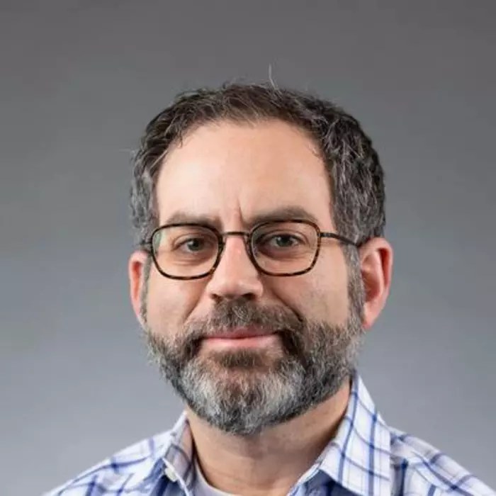 A white man with greying hair, who wears glasses and a white and blue checked shirt