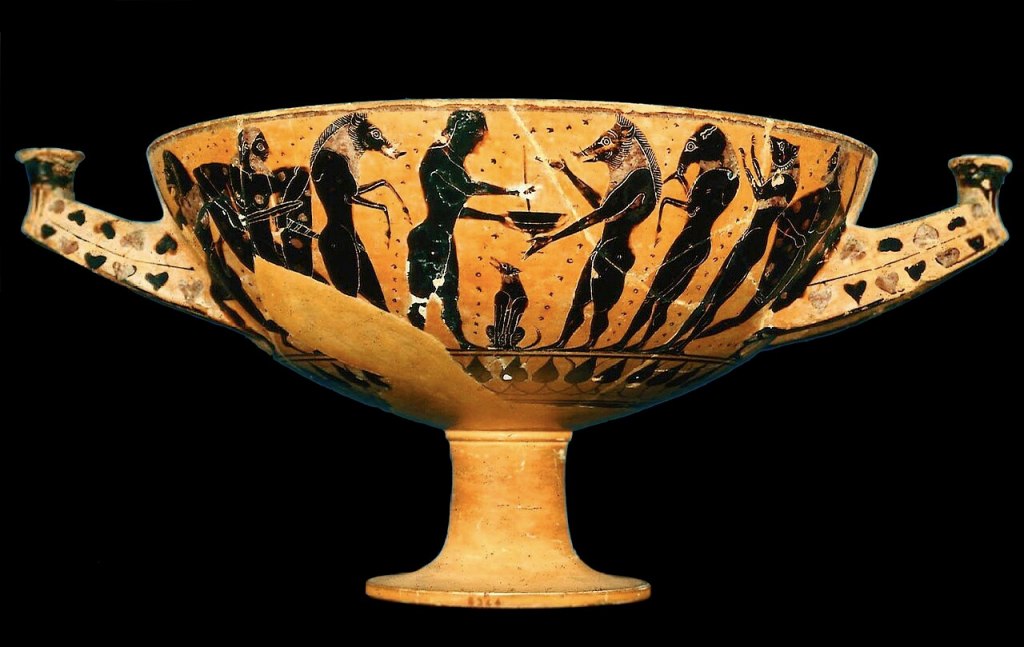 Image of an ancient Greek drinking cup with black background. The drinking cup has a long stem and handles on either side. The painting on the outside of the cup shows men with animal heads lining up in front of a naked man (Odysseus), who holds a cup out to them. They line up both in front of and behind Odysseus. All are painted black with incised details. A dog sits under Odysseus’ outstretched hand holding the cup. A frieze of palm leaves runs below, and heart-shaped leaves decorate the handles.